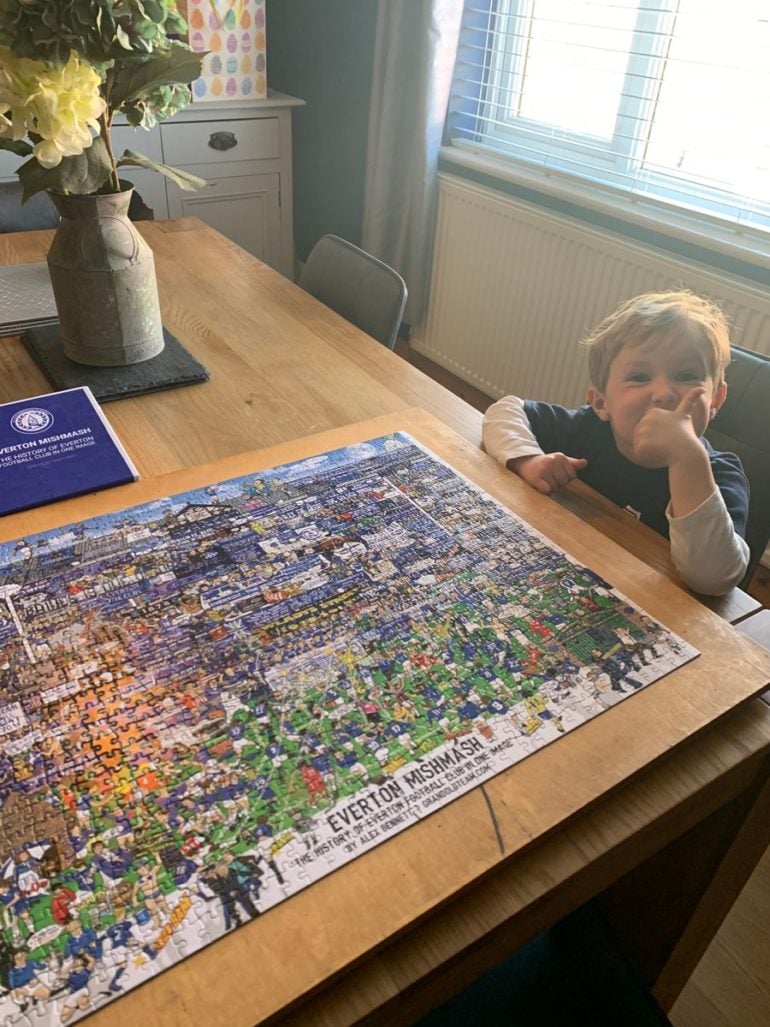 The image shows a young child sitting at a wooden dining table, resting their chin on their hand while looking at a completed jigsaw puzzle. The puzzle is titled "Everton Mishmash: The History of Everton Football Club in One Image" by Alex Bennett, featuring a colorful and detailed illustration of various scenes, players, and elements related to Everton FC, predominantly in blue and green hues. The puzzle box is also on the table to the left, displaying the same design and title. A vase with dried yellow flowers sits on a black coaster next to the box. In the background, there is a white cabinet with a decorative piece featuring colorful polka dots, and a large window with blue blinds and a radiator beneath it, letting in natural light. The room has a cozy, domestic feel with light-colored walls and a relaxed atmosphere.