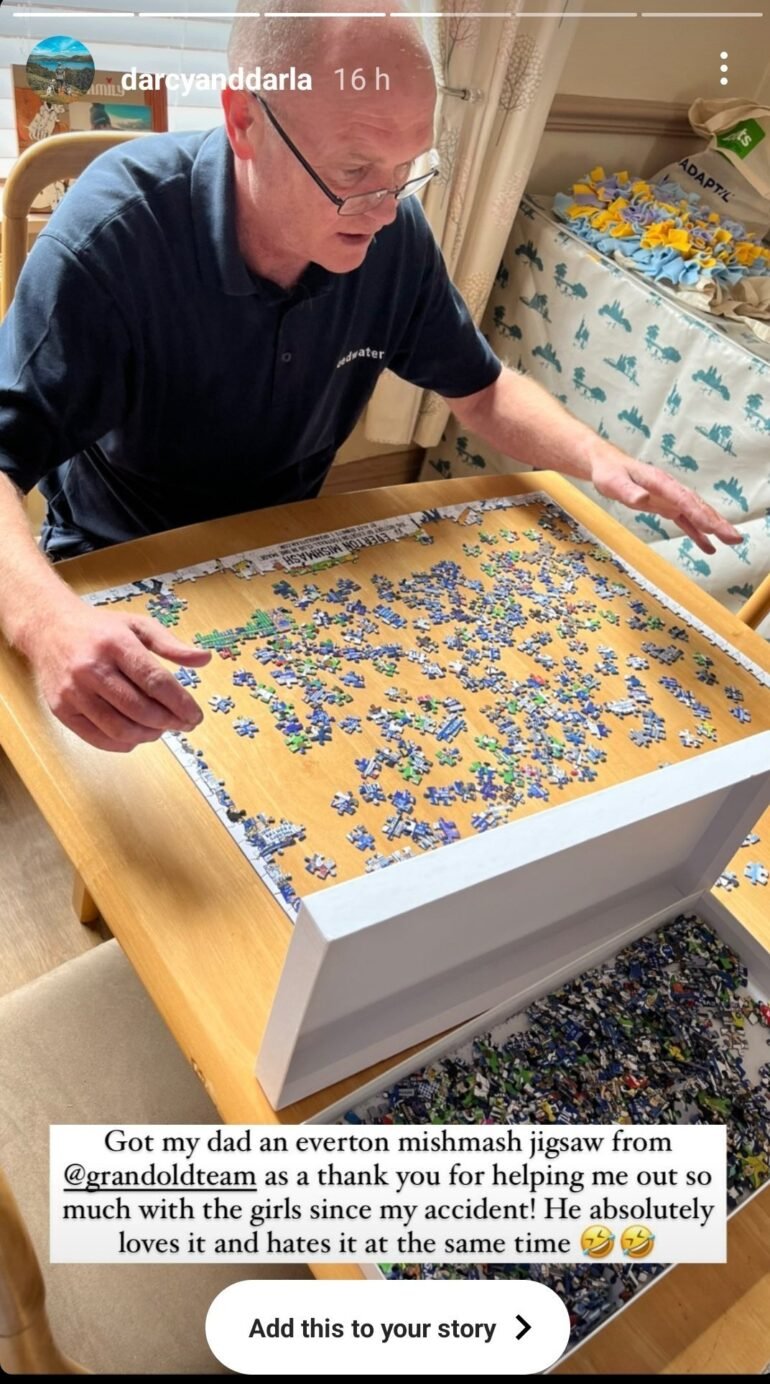 A man with glasses and a navy polo shirt is working on a jigsaw puzzle at a wooden dining table. He appears focused as he carefully selects a piece from the puzzle box. The puzzle is the Everton Mishmash, a detailed illustration of Everton Football Club's history. The edges of the puzzle are mostly completed, while the centre remains a mix of unplaced pieces. A caption overlay from an Instagram story mentions that the puzzle was a gift from the user's account (@grandoldteam) to their father as a thank-you. The background shows a cushioned bench with a blanket and a shopping bag on it.