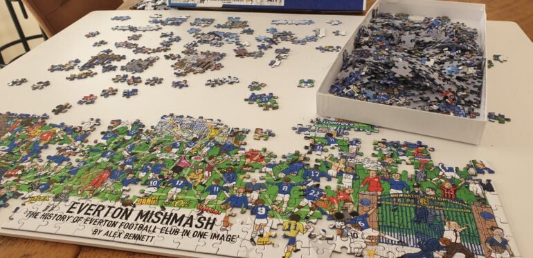 A partially completed Everton Mishmash jigsaw puzzle is laid out on a white table. The lower section, filled with colourful cartoon-style illustrations of famous Everton players and moments, is mostly assembled, while the upper portion still has many missing pieces. Loose puzzle pieces are scattered around, and a large box filled with unplaced pieces sits on the table. The puzzle features bright blues, greens, and reds, with Everton-related text and imagery.